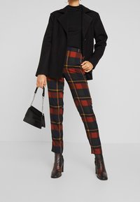 Person som bär en svart blazer, svart topp, rutig byxa i röd, marinblå och senapsgul, håller en liten svart handväska och har på sig svarta klackstövlar.