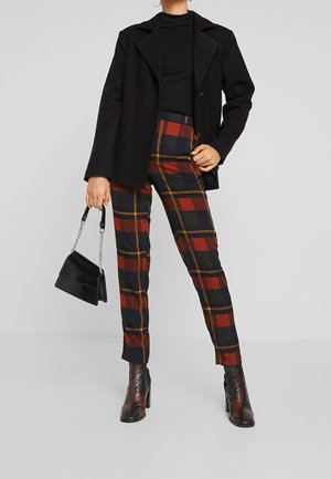 Personne portant un blazer noir, un haut noir et un pantalon à carreaux dans des teintes de rouge, marine et moutarde, tenant un petit sac à main noir et portant des bottines à talons noires.