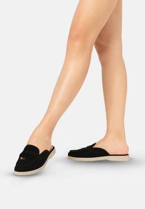 Benen met zwarte slip-on loafers met beige zolen, één voet licht opgetild, tegen een effen lichte achtergrond.