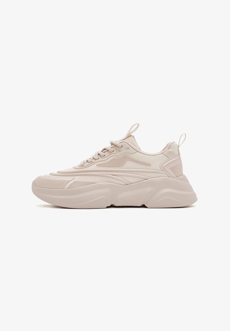 Zapatilla beige con parte superior de malla texturizada, suela de goma sólida y diseño curvado. Presenta cordones, lengüeta de agarre y franjas laterales distintivas.