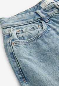 Ljust blå denimjeans med en sliten look, föreslagna med knappstängning, metallnitar och detaljerad sömnad på fickor och sömmar.