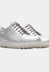 Silverfärgade skor med texturerad yta, rund tå och grå snörning. Har en tjock vit sula med cirkulära grepp och röda varumärkeslogotypdetaljer.