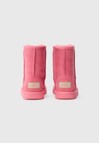 Bottines en daim rose avec doublure en fourrure, semelles en caoutchouc rose plates et logo UGG sur une étiquette beige à l'arrière. Texture unie, sans motifs.