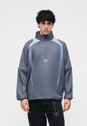 Mladý muž oblečený v voľnom sivom pulóvri so svetlomodrými panelmi a polovičným zipsom na golieri, v kombinácii s čiernymi nohavicami so štýlovými malými bielymi cvočkami.