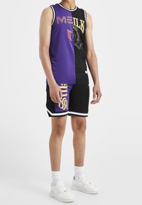 Fioletowy i czarny sportowy tank top z graficznymi wzorami, w połączeniu z czarnymi szortami z fioletowymi akcentami i żółtym napisem, uzupełniony białymi trampkami.