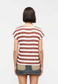 Γυναίκα με ίσια σκούρα μαλλιά, που φοράει ριγέ τοπ κοντών μανικιών σε χρώματα σκουριάς και λευκού και μπλε τζιν μίνι φούστα με μπεζ τσέπες, στέκεται γυρισμένη προς το πίσω μέρος.