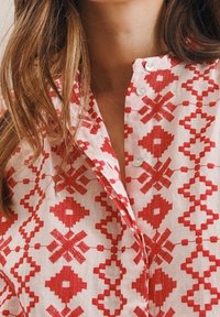 Košile červené a bílé barvy s geometrickými vzory, knoflíky a lehkým, texturovaným materiálem. Detailní záběr oblasti límce.