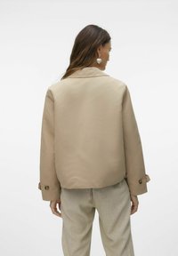 Beige, siled tekstuuriga jakk kraega ja nööpidega mansettidega, kombineeritud heledate, mustriliste pükstega. Minimalistlik disain.