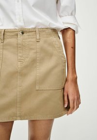 Khaki denimhõlmik, millel on vormiv vöökoht, eesmine tasku ja hemliin, mis on just üle põlve. Materjal on siledat tekstuuri ja peenet sära.