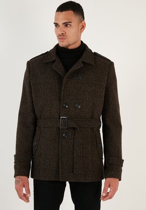 Brunmønstret dobbeltbrystet frakke med bælte, der har store knapper, krave og sidelommer, båret over en sort turtleneck.
