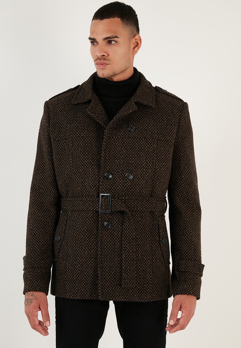 Brauner gemusterter Doppelbreasted-Mantel mit einem Gürtel, großen Knöpfen, einem Kragen und Seitentaschen, getragen über einem schwarzen Rollkragenpullover.