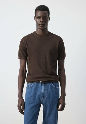 Homme portant un pull à manches courtes marron foncé et un jean bleu, debout devant un fond gris clair uni.