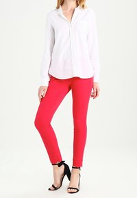 Femme portant une chemise blanche à manches longues avec des boutons, un pantalon ajusté rose vif avec des ourlets festonnés, et des escarpins noirs à bride cheville.