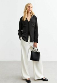 Zwarte blouse met lange mouwen en kraag, voorzien van twee zakken, gecombineerd met witte wijde broekspijpen. Een kleine zwarte tas met handvat maakt de look af.