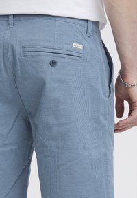 Light blue textured pants with a subtle pattern, featuring a back pocket with a button and a label on the waistband. Side pocket visible.
