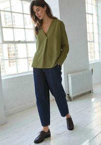 Blouse vert olive avec un décolleté en V et une coupe décontractée, associée à un pantalon en denim bleu foncé. Le mannequin porte des chaussures à lacets brun foncé.