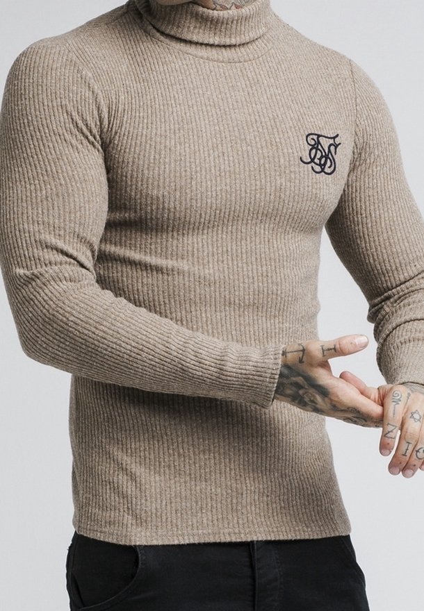 Pull à col roulé beige côtelé avec logo noir brodé sur la poitrine, porté par une personne tatouée ajustant sa manche.
