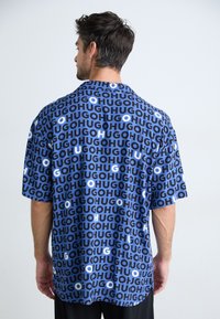 Short-sleeve shirt featuring a blue fabric with a bold repeating 'HUGO' print in white and varied shades of blue. Classic collar design.