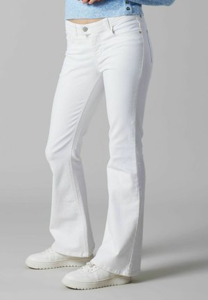 Kvinde iført hvide vidde jeans og hvide sneakers, stående mod en ensfarvet lys grå baggrund.