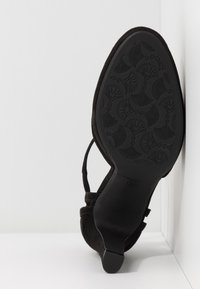 Semelle d'une chaussure à talon haut noire avec un motif floral sur la bande de roulement, placée à la verticale contre un coin blanc de mur et de sol.