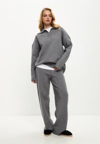 Šedý svetr s límcem a volným střihem, doplněný o široké kalhoty ve stejném stylu. Měkká textura, minimalistický design a kontrastní černé boty.