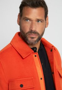 Oranges Wollmischjacke mit einem abgerundeten Kragen, Vorderknopfschluss und zwei Brusttaschen. Mit kontrastierenden orangen Seiten und strukturiertem Stoff.