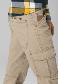 Beige Cargo-Hosen mit mehreren Taschen, verstärkten Nähten und einer glatten Textur. Verfügt über ein Lederetikett auf der Rückseite und Knopfverschlüsse.