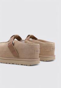 Pantofle in pelle scamosciata beige con una texture liscia, dotate di un'etichetta con il marchio sul lato e una suola in gomma beige con un motivo a rilievo.