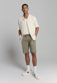 Lichtbeige overhemd met korte mouwen, witte tanktop, olijfgroene shorts, witte sokken en crèmekleurige sportschoenen met blauwe accenten.