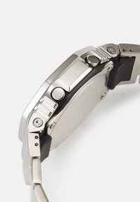 Relógio em aço inoxidável com acabamento escovado, caixa redonda, múltiplos botões e uma bracelete de borracha com detalhes de hardware visíveis.