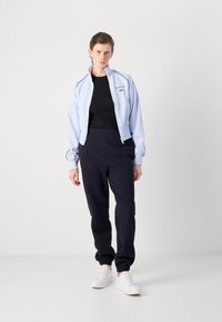Svetlomodrá bunda na zips s čiernymi doplnkami, čierne tričko a námořnícké tepláky. Model v bielych teniskách, stojaci na neutrálnom pozadí.