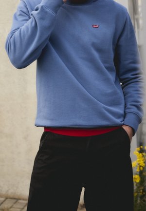 Person, die einen blauen Levi’s Sweatshirt über einem roten Shirt und schwarzen Hosen trägt, steht mit einer Hand in der Tasche und der anderen nahe am Gesicht.