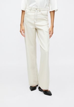 Woman wearing high-waist, wide-leg cream-colored jeans paired with black pointed shoes and a white top, standing against a white background.