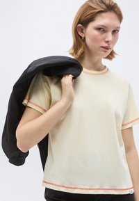 Νεαρό άτομο με ανοιχτόχρωμα μαλλιά φοράει μια κρεμ κοντομάνικη μπλούζα με κόκκινη ραφή, κρατώντας ένα μαύρο μπουφάν στον ώμο του πάνω σε απλό φόντο.