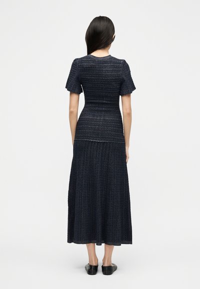 Γυναίκα που στέκεται με την πλάτη στην κάμερα, φορώντας ένα μπλε σκούρο υφασμάτινο πλεκτό φόρεμα με κοντά μανίκια και μαύρα επίπεδα παπούτσια.