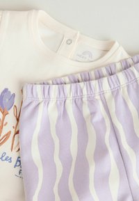 Babaöltözék krémszínű pamut pólóval, virágmintával és levendulás csíkos nadrággal, rugalmas derékrésszel. Puha tapintású, kényelmes szabású.