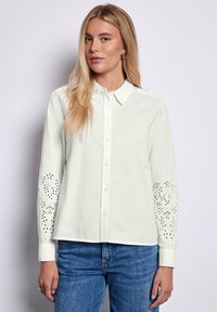 Camisa blanca de botones con mangas largas que presenta patrones florales recortados en las mangas, hecha de suave tela de algodón. Combinada con jeans azules.