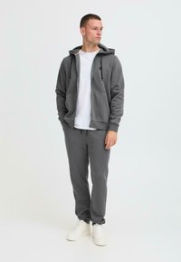 Grå bomullshoodie med frontglidelås, frontlommer og matchende joggebukser. Brukt med en hvit t-skjorte og hvite joggesko.