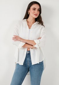 Bílá, lehká košile na knoflíky s texturovanou tkaninou, dlouhými rukávy ohrnutými na manžetách, kombinovaná s modrými džíny s vysokým pasem.