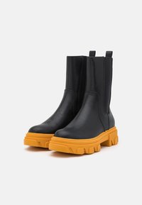 Bottes chevilles en cuir noir avec des semelles en caoutchouc jaunes vives et des panneaux élastiques sur les côtés, présentées sur un fond blanc.