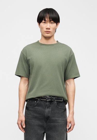 Mladý muž s rovnými čiernymi vlasmi, oblečený v voľnom olivovo zelenom tričku a vyblednutých čiernych džínsoch s čiernym opaskom s cvočkami.