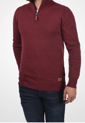 Mann trägt einen bordeauxroten Pullover mit Viertel-Reißverschluss, gerippten Bündchen und Saum, kombiniert mit dunkelblauen Jeans, vor einfachem weißem Hintergrund.