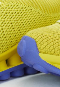Φωτεινά κίτρινα αθλητικά παπούτσια με υφασμάτινο άνω μέρος με υφή και μωβ καουτσούκ σόλα, με σαφή στοιχεία απορρόφησης κραδασμών.