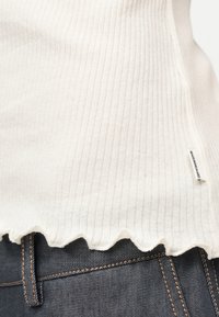 Κρεμ ριμπ πλεκτό τοπ με βολάν στο τελείωμα και μικρή πλαϊνή ετικέτα, φορεμένο πάνω από σκούρο τζιν με ραφές σε αντίθεση.