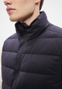 Melnā izolētā veste ar augstu apkakli, ar izšūtu rakstu un uz krūšu zonas izspiedumu ar zīmola nosaukumu "BOSS".