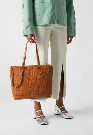 Borsa a tracolla in pelle color cuoio con logo in rilievo, caratterizzata da un accentuazione a catena. Ha due manici e una forma strutturata. Indossata con pantaloni chiari.