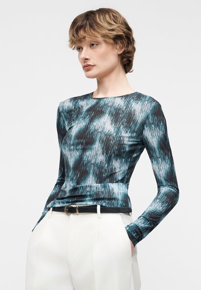 Άτομο που φοράει μπλούζα με μακριά μανίκια, σε μπλε και μαύρο αφηρημένο σχέδιο, η οποία είναι μισοκουμπωμένη μέσα σε ψηλόμεσα λευκά παντελόνια με μαύρη ζώνη.