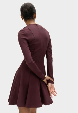 Kvinde med mørkt hår i en knold iført en langærmet, burgunderfarvet kjole med tætsiddende krop og udvidet nederdel, stående med hænderne foldet bag ryggen.