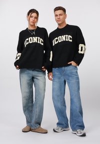 Black knit sweaters with “ICONIC” in cream, paired with light blue jeans. One model wears beige shoes, the other gray sneakers.