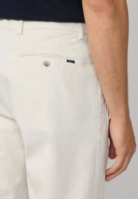 Uski blizu prikaz osobe koja nosi off-white hlače s dugmetom na stražnjem džepu i malom Hackett oznakom, ruka opuštena uz tijelo.